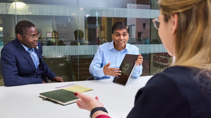 Three smartly dressed people sat round an office table in conversation, one of them is holding a tablet device. Two notepads can be seen on the table.