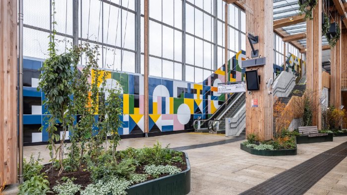 The interior of the London Brent Cross West station. Large wooden pillars, plant containers and an escalator can be seen along the station entrance.