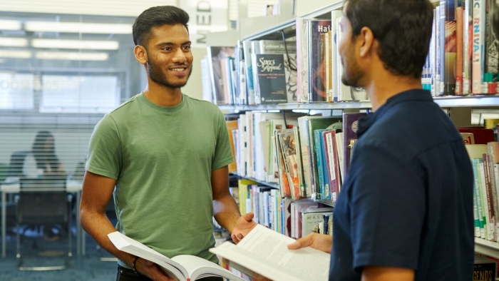 Two casually dressed students stood in a library in conversation. Both are holding open books.